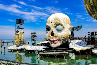 A dramatic outdoor stage setup on water, featuring a massive, artistic sculpture of a face with a painted smile and one eye covered. The stage appears to have walkways and platforms, with additional mechanical structures and set pieces around. The backdrop is a clear blue sky and calm waters.