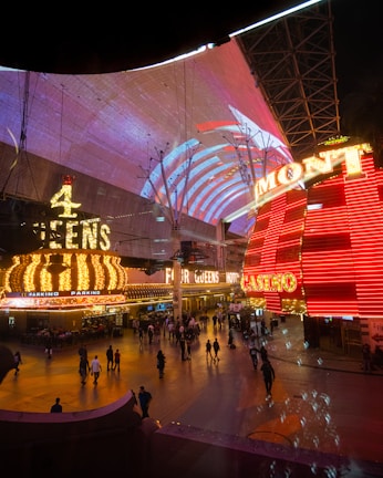A bustling nighttime scene with illuminated casino signage towering over a busy pedestrian street. Neon lights in red and yellow highlight the names of various establishments. People are walking in different directions, enjoying the vibrant atmosphere.