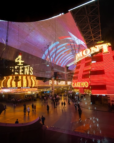 A bustling nighttime scene with illuminated casino signage towering over a busy pedestrian street. Neon lights in red and yellow highlight the names of various establishments. People are walking in different directions, enjoying the vibrant atmosphere.