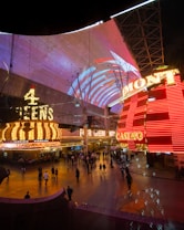 A bustling nighttime scene with illuminated casino signage towering over a busy pedestrian street. Neon lights in red and yellow highlight the names of various establishments. People are walking in different directions, enjoying the vibrant atmosphere.