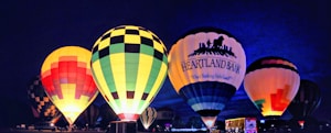 A vibrant display of hot air balloons illuminated against a night sky, featuring various patterns and colors. The balloons are arranged in a row, with some displaying brand logos. Spectators and vehicles can be seen at the base of the balloons, adding to the lively atmosphere.
