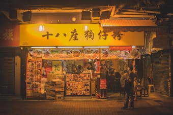 An inviting street restaurant with a bright yellow signboard displaying text in a foreign language. The restaurant's facade is decorated with various food pictures and colorful advertisements. Inside, people are visible, sitting and standing, creating a lively and bustling atmosphere. The scene is warmly lit, suggesting nightlife or evening eating culture.