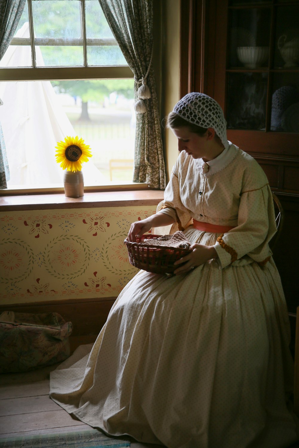 woman wearing white long-sleeved dress sitting and holding wicker basket near window
