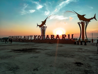 A coastal area at sunset featuring large letters spelling out 'Siringlaut Kotabaru' with two decorative swordfish statues emitting water streams on each side. The sun is partially obscured by the letters, casting a warm glow over the scene, with a mix of clouds in the sky and a few people walking on the pavement.