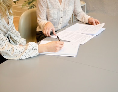 woman signing on white printer paper beside woman about to touch the documents