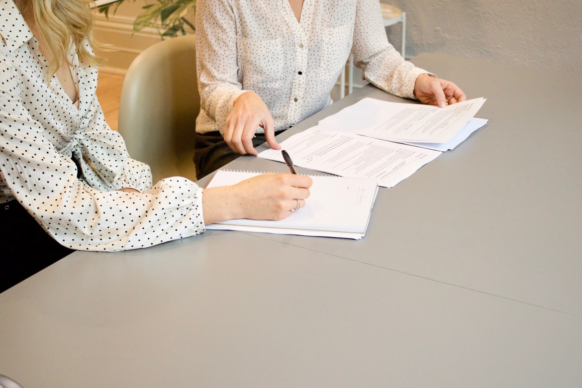  a person writing something on a document with a pen while sitting next to a person who is flipping through documents at a table
