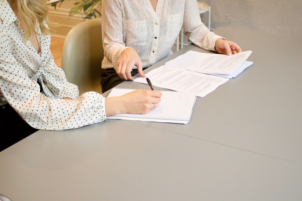 woman signing on white printer paper beside woman about to touch the documentsby Gabrielle Henderson