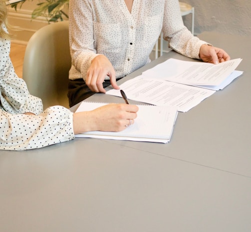 woman signing on white printer paper beside woman about to touch the documents