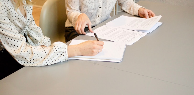 woman signing on white printer paper beside woman about to touch the documents