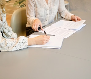 woman signing on white printer paper beside woman about to touch the documents