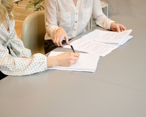 woman signing on white printer paper beside woman about to touch the documents