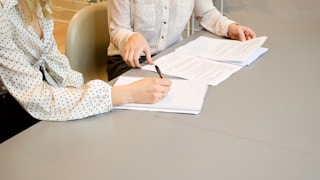 woman signing on white printer paper beside woman about to touch the documents