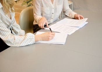 woman signing on white printer paper beside woman about to touch the documents