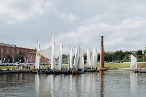 A series of traditional wooden boats with large white sails are lined up along a calm body of water. The boats are moored near a brick industrial building, with a tall chimney and a small group of people gathered nearby. Flags with various colors decorate the boats. Lush greenery and trees are visible in the background under a partly cloudy sky.