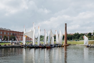 A series of traditional wooden boats with large white sails are lined up along a calm body of water. The boats are moored near a brick industrial building, with a tall chimney and a small group of people gathered nearby. Flags with various colors decorate the boats. Lush greenery and trees are visible in the background under a partly cloudy sky.