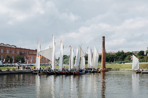A series of traditional wooden boats with large white sails are lined up along a calm body of water. The boats are moored near a brick industrial building, with a tall chimney and a small group of people gathered nearby. Flags with various colors decorate the boats. Lush greenery and trees are visible in the background under a partly cloudy sky.
