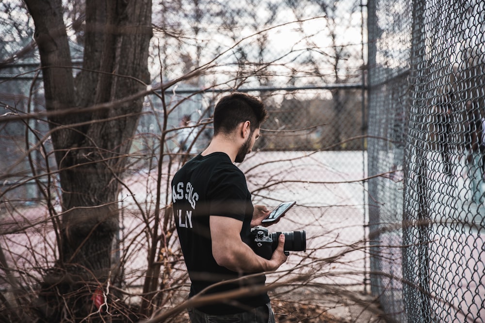 man using phone and holding camera standing in the corner fence