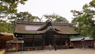 A traditional Japanese shrine with a thatched roof and wooden structure sits surrounded by lush greenery. The entrance has large wooden doors, and the complex seems historical and well-preserved. Two people are seen walking towards the shrine, adding a sense of scale. Various trees are visible in the background, suggesting a peaceful, natural setting.