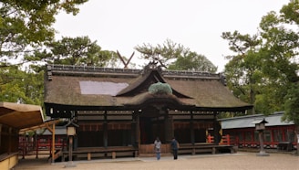 A traditional Japanese shrine with a thatched roof and wooden structure sits surrounded by lush greenery. The entrance has large wooden doors, and the complex seems historical and well-preserved. Two people are seen walking towards the shrine, adding a sense of scale. Various trees are visible in the background, suggesting a peaceful, natural setting.
