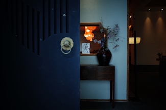 A dimly lit interior space featuring a large dark door with a round metallic handle. Behind the door, a wooden table holds a black vase with branches and flowers. A wall-mounted mirror reflects warm, hanging lights, adding a cozy glow to the scene.