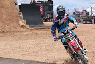 A motocross rider wearing a colorful blue and red outfit, complete with protective gear and a helmet, maneuvers a red dirt bike on a track. The background includes a large dirt ramp and barriers, with banners and a partially cloudy sky overhead.