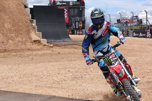 A motocross rider wearing a colorful blue and red outfit, complete with protective gear and a helmet, maneuvers a red dirt bike on a track. The background includes a large dirt ramp and barriers, with banners and a partially cloudy sky overhead.