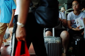 Several people are seated in a public transit setting, possibly a bus or train. One person is holding a book, another person is standing, and there are suitcases and bags around them. The individuals appear to be engaged in their own activities.