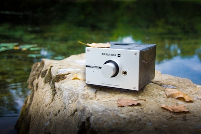 A metal audio device with a large knob and labeled 'ERZETICH' is placed on a rock near a body of water. The device has a rectangular shape and appears slightly worn. Fallen leaves are scattered on the rock around the device. The water in the background is calm, reflecting the greenery of nearby trees.