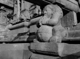 A stone sculpture of a mythological figure with folded hands is prominently featured. The figure has an intricate design and is set against a backdrop of detailed architectural stonework, indicative of ancient craftsmanship.