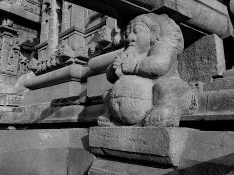 A stone sculpture of a mythological figure with folded hands is prominently featured. The figure has an intricate design and is set against a backdrop of detailed architectural stonework, indicative of ancient craftsmanship.