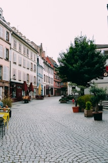 A quaint European street features cobblestone pavement lined with charming buildings on one side. The buildings exhibit a variety of pastel colors, adding to the rustic atmosphere. Outdoor seating with tables and chairs is present, along with a few people relaxing. Large planters and trees contribute to a serene and inviting environment.