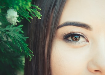 A close-up of a woman's face, focusing on her eye and eyebrow. The eye captures a reflection and is adorned with subtle makeup. To the left, green foliage with detailed leaves is visible, adding a natural element.