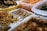 An assortment of prepared foods is organized in transparent plastic containers. The foods appear to be various types of seasoned or pickled dishes, possibly including kimchi, greens, and fried items. A yellow basket holding a variety of fried snacks is visible in the background.