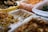 An assortment of prepared foods is organized in transparent plastic containers. The foods appear to be various types of seasoned or pickled dishes, possibly including kimchi, greens, and fried items. A yellow basket holding a variety of fried snacks is visible in the background.