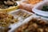 An assortment of prepared foods is organized in transparent plastic containers. The foods appear to be various types of seasoned or pickled dishes, possibly including kimchi, greens, and fried items. A yellow basket holding a variety of fried snacks is visible in the background.