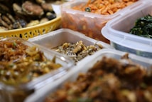 An assortment of prepared foods is organized in transparent plastic containers. The foods appear to be various types of seasoned or pickled dishes, possibly including kimchi, greens, and fried items. A yellow basket holding a variety of fried snacks is visible in the background.