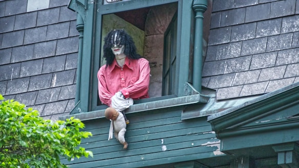 A mannequin with a pale face, black curly hair, and a red shirt leans out of a window on a weathered building with green trim and dark shingles. The mannequin holds a doll with brown hair.