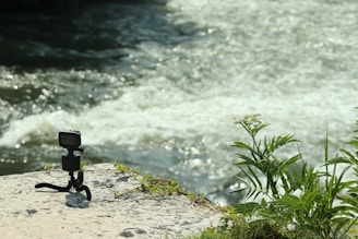 A small action camera is mounted on a flexible tripod on a rock beside a flowing river. The scene includes some green plants growing nearby, and sunlight reflecting off the water creates a bright, natural setting.