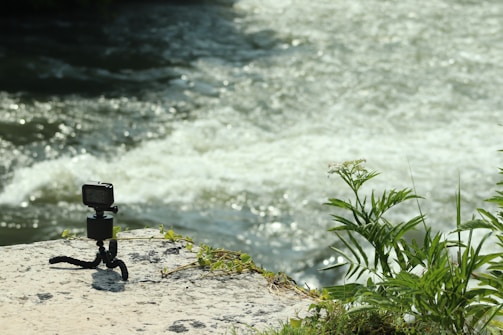 A small action camera is mounted on a flexible tripod on a rock beside a flowing river. The scene includes some green plants growing nearby, and sunlight reflecting off the water creates a bright, natural setting.