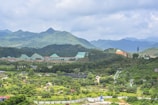 A vast landscape featuring lush green hills and mountains under a slightly cloudy sky. The foreground includes various greenery and trees with pathways and structures. There are visible human-made elements like a building with a sloped roof and some wind turbines.