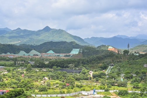 A vast landscape featuring lush green hills and mountains under a slightly cloudy sky. The foreground includes various greenery and trees with pathways and structures. There are visible human-made elements like a building with a sloped roof and some wind turbines.