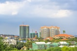 A cityscape featuring several high-rise buildings under construction and completed structures. The skyline includes cranes and scaffolding, indicating ongoing development. The area is surrounded by greenery with trees and some residential buildings.