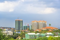 A cityscape featuring several high-rise buildings under construction and completed structures. The skyline includes cranes and scaffolding, indicating ongoing development. The area is surrounded by greenery with trees and some residential buildings.