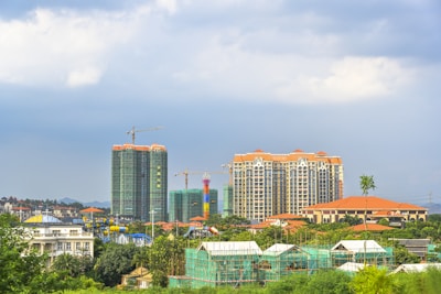 A cityscape featuring several high-rise buildings under construction and completed structures. The skyline includes cranes and scaffolding, indicating ongoing development. The area is surrounded by greenery with trees and some residential buildings.