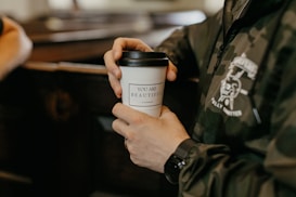 A person holds a takeaway coffee cup with a message printed on it that reads 'YOU ARE BEAUTIFUL'. They are wearing a dark camouflage jacket with a design on it. The focus is on the hands and the cup, with a blurred background.