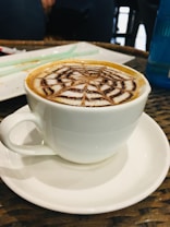 A white cup filled with cappuccino or latte featuring an intricate foam art design resembling a spiderweb. The cup sits on a matching white saucer placed on a wicker table. In the background, blurred objects and a blue bottle are visible, creating a cozy cafeteria environment.