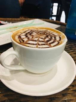 A white cup filled with cappuccino or latte featuring an intricate foam art design resembling a spiderweb. The cup sits on a matching white saucer placed on a wicker table. In the background, blurred objects and a blue bottle are visible, creating a cozy cafeteria environment.