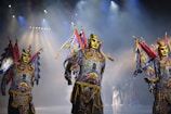 Performers in ornate costumes with gold masks and colorful, elaborate outfits stand on a stage under dramatic lighting. The costumes include bright blues, reds, and yellows with intricate patterns, and the figures appear to be part of a theatrical or cultural performance.