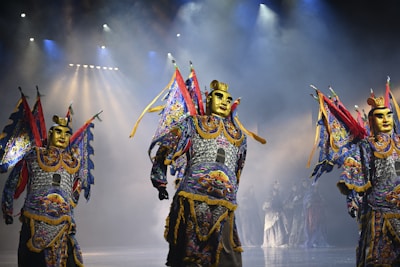 Performers in ornate costumes with gold masks and colorful, elaborate outfits stand on a stage under dramatic lighting. The costumes include bright blues, reds, and yellows with intricate patterns, and the figures appear to be part of a theatrical or cultural performance.