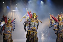 Performers in ornate costumes with gold masks and colorful, elaborate outfits stand on a stage under dramatic lighting. The costumes include bright blues, reds, and yellows with intricate patterns, and the figures appear to be part of a theatrical or cultural performance.